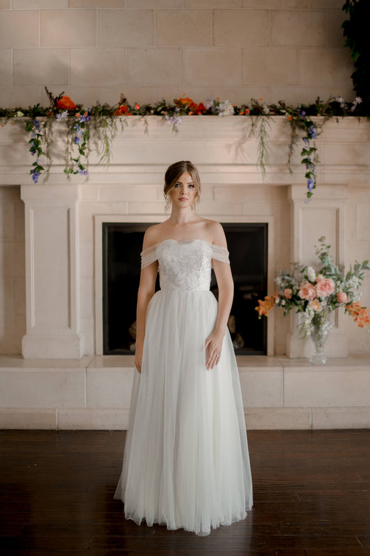 A styled look of the penelope gown on a bride; the Penelope A-line gown has an off-the-shoulder arm drape and gathered glitter tullé skirt. 
