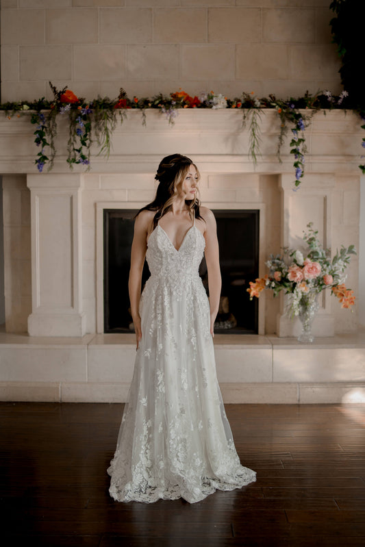 A full length shot of the Wilhelmina gown from the front on a bride; it shows off the deep v neckline, beaded floral overlay and romantic a-line silhouette. 