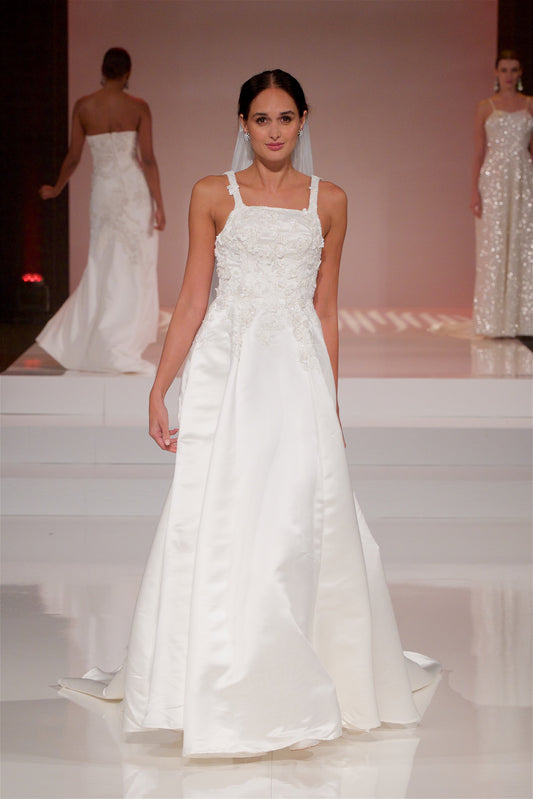 A full front  view  the sabina gown on a runway, the beaded 3d lace bodice and satin skirt shine in the light. The gown is paired with the Cara veil but it just pokes out behind her head. 