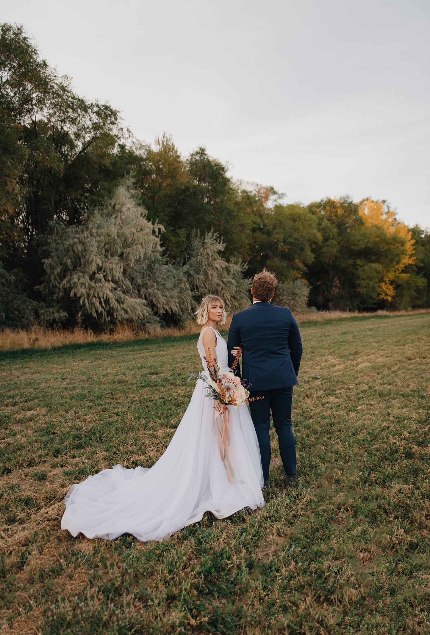 A styled shoot of a bride wearing the persephone gown, without a petticoat, standing with her groom and looking back at the camera. The long tulle train extends out. 