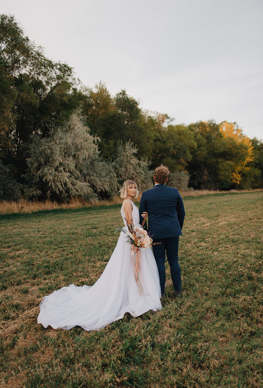 A styled shoot of a bride wearing the persephone gown, without a petticoat, standing with her groom and looking back at the camera. The long tulle train extends out. 